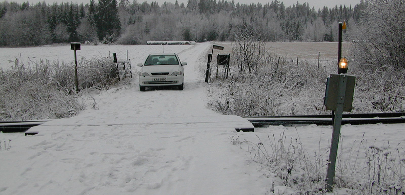 Planovergang med enkel varsellampe og bil på snødekt vei i landlig vinterlandskap.