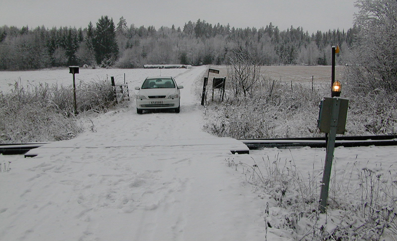 Planovergang med enkel varsellampe og bil på snødekt vei i landlig vinterlandskap.