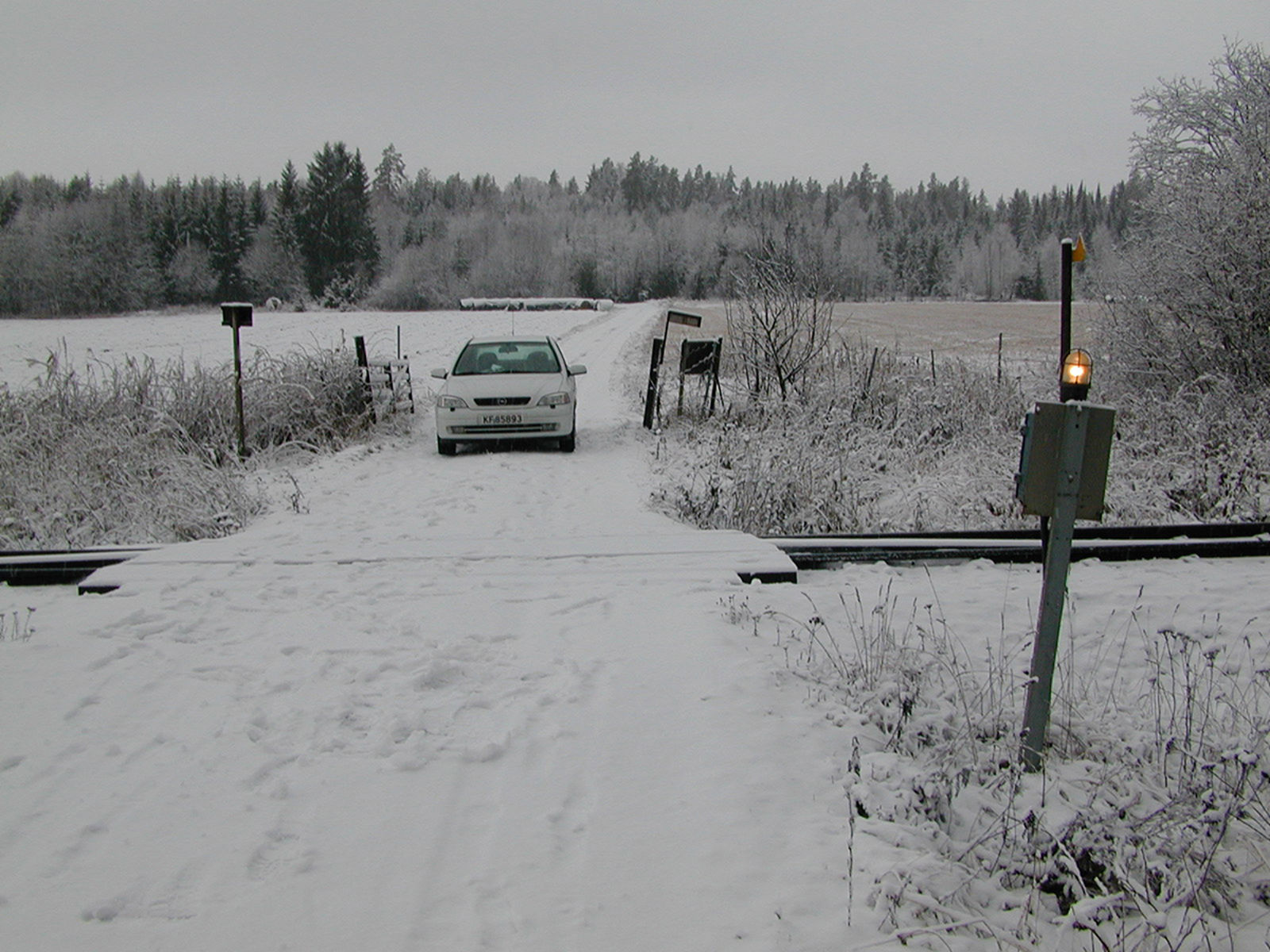 Planovergang med enkel varsellampe og bil på snødekt vei i landlig vinterlandskap.