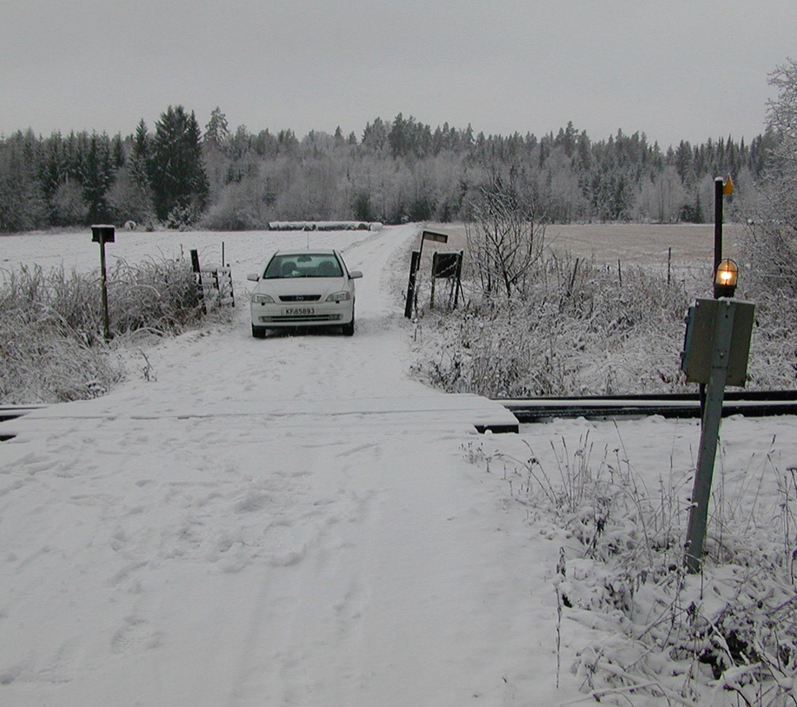 Planovergang med enkel varsellampe og bil på snødekt vei i landlig vinterlandskap.