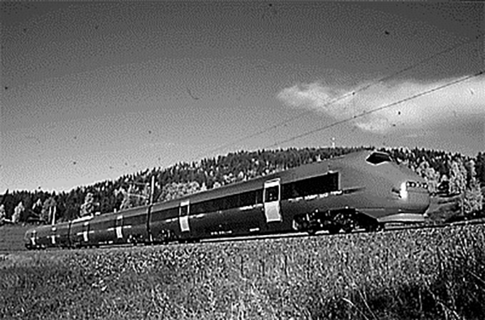 Flytoget type BM71 på høyhastighetsbane i kupert landskap med skog i bakgrunnen.